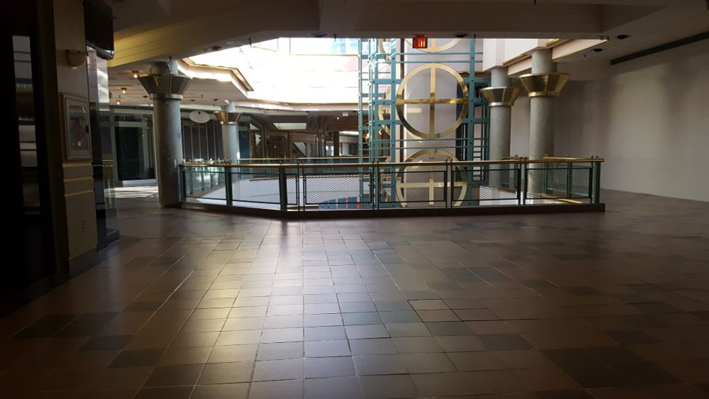 a dark area of  mall. there's light spilling through a skylight towards the far end of the hall where an elevator shaft with large brass circles sits.