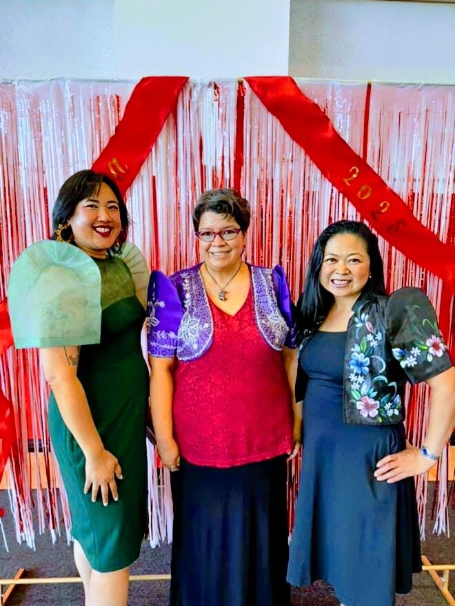 Three filipinas standing side by side, smiling at the camera. They are dressed in vibrant, formal attire with traditional elements (all three wearing Filipiniana). The woman on the left wears a dark green dress with sheer green fabric —butterfly sleeves— draped over her shoulders. The woman in the middle is dressed in a dark magenta top with intricate white lace patterns and a violet butterfly-sleeved bolero with embroidered details. The woman on the right wears a navy blue dress with a butterfly-sleeved, floral embroidered bolero.

The background features a decorative backdrop of vertical red and white fringe curtains. Red banners with gold lettering drape diagonally above them.