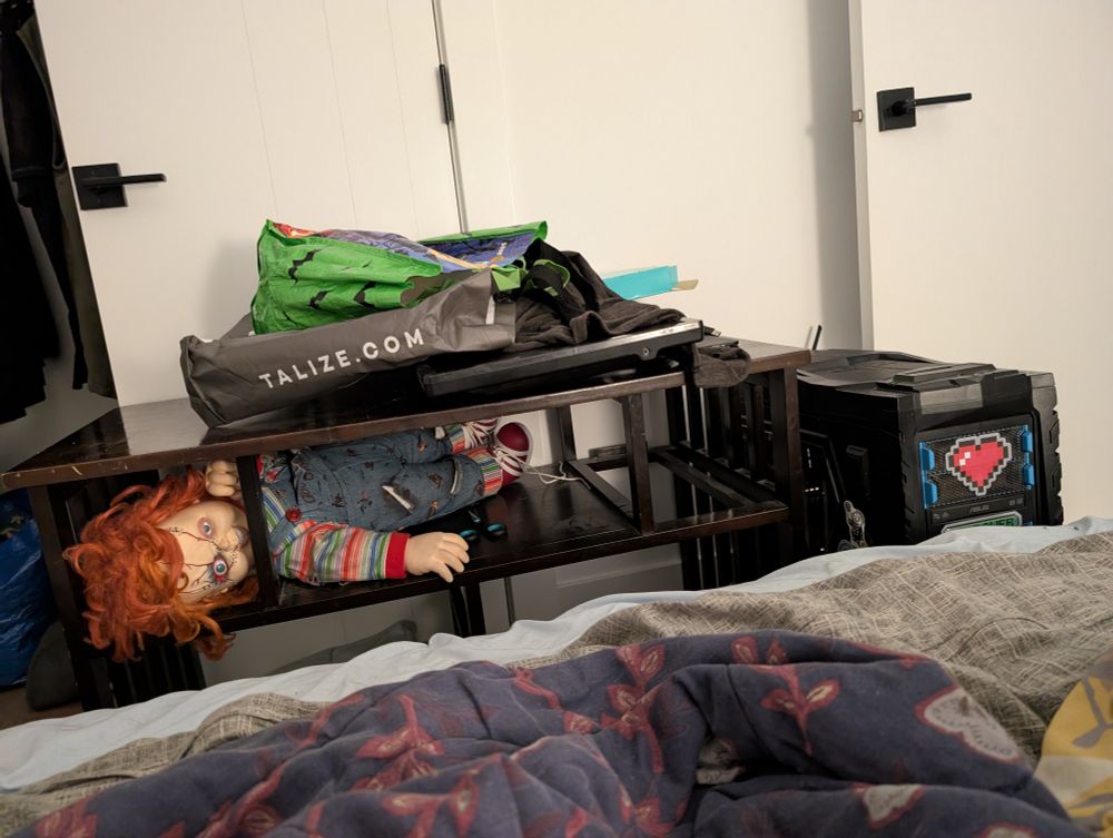 A photo of my computer partially set up at the foot of my bed on a temporary "desk". Chucky from Child's Play is hanging out sideways on the shelf. Like a creep. 