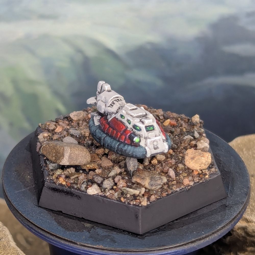 A miniature of a Savannah Master hovercraft for Battletech, painted with a dark skirt, red side panels, and white down the center. 