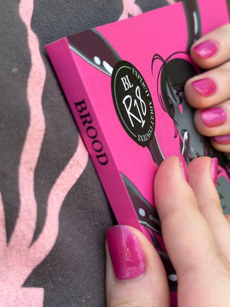 close-up photo of a copy of a book called "BROOD". the cover is mostly hidden but there's a character with a tentacle in their mouth and a BL / R18 warning on the front. the cover is pink.