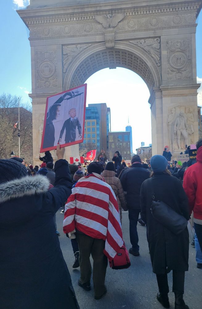 A woman holds a sign with a collage of Elon Musk holding the puppet strings for a President Trump marionette.