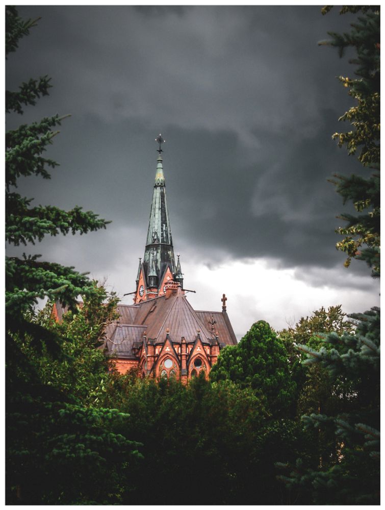 Photo: an old church stands tall, peeking through dark evergreens. The orange walls and steep rust colored roof contrast with the deep green foliage. The spire, covered in pale green tiles, reaches into a stormy sky. 