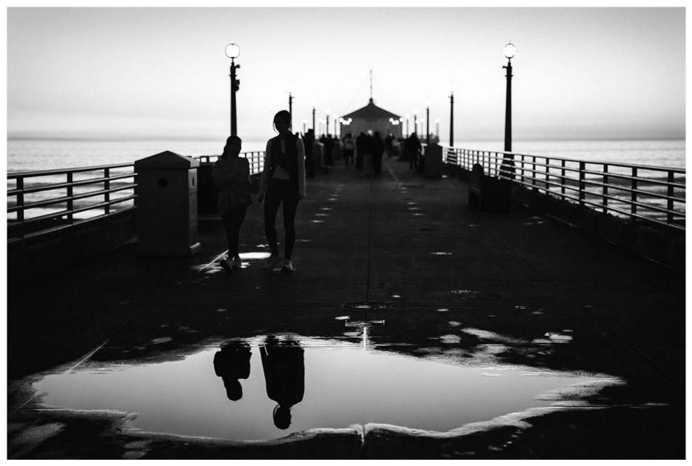 Black and white photo looking down the pier in Manhattan Beach. The twilight sky is soft and muted, reflecting off the ocean in the distance and off of a large puddle in the foreground. Streetlamps line the walkway as people wander back from watching the sunset. Two women walk and talk, their silhouettes reflected in the puddle. 
