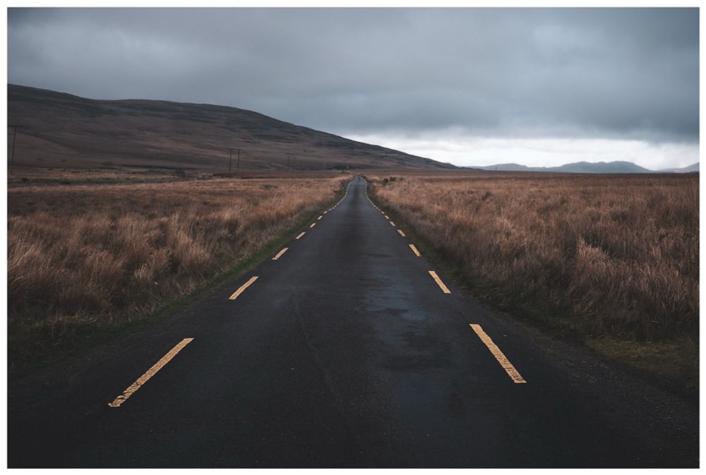 Photo: staring down a long, narrow road toward the mountains at the horizon. Open fields of wheat-colored grass stretch out on either side and storm clouds gather in the distance. 