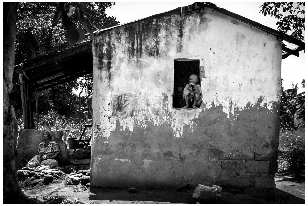 Black and white photo. A boy crouches, pensive, in the solitary open window of a small farmhouse. The walls bow slightly and are in need of replastering. A wooden overhang extends to the left of the house, providing shade for the boy’s mother, who is resting in the afternoon heat. Banana palms grow in the surrounding fields. 