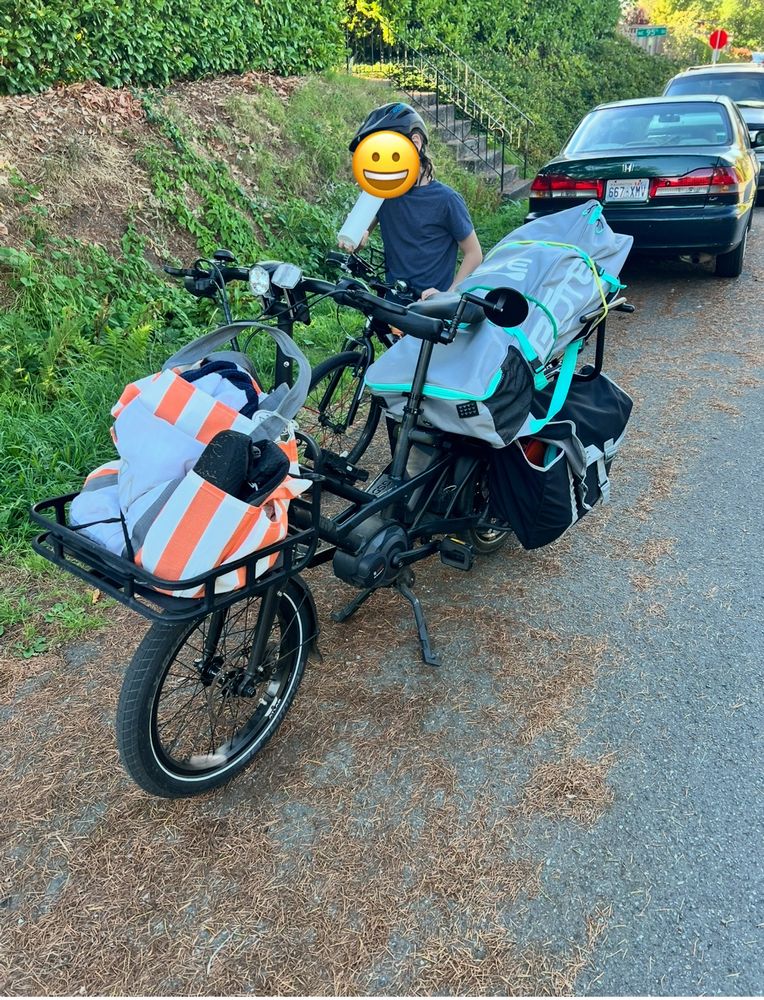 Cargo bike carrying inflatable paddle board and beach bag, child on second bike behind it. 