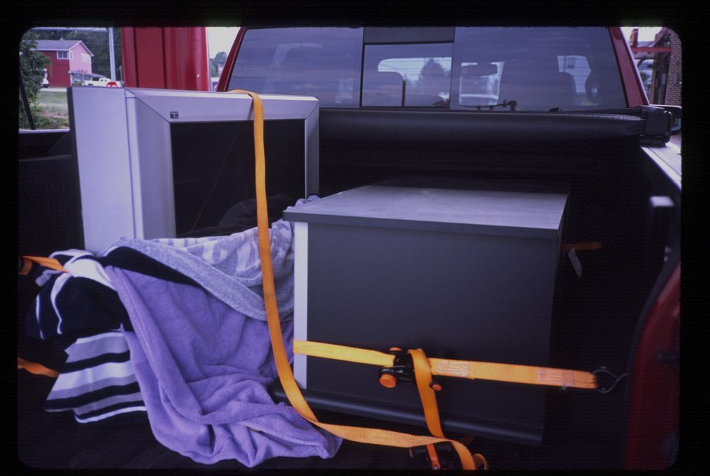 a large (30”) CRT sits in the back of a red truck, along with its stand. Both date from 2004 or so, and are secured with orange ratchet straps.