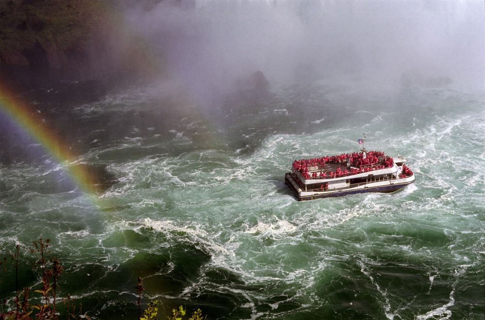 A photo of a the "Niagara Thunder" sightseeing boat heading towards Niagara Falls. Mist is starting to encroach on the boat and it's passengers at the top of the frame, while a rainbow (plus a second faint bow) arches behind them. The water is a deep black/blue where it isn't white from turbulence. All the people on the deck of the ship are wearing red ponchos, save for one at the stern in blue. 