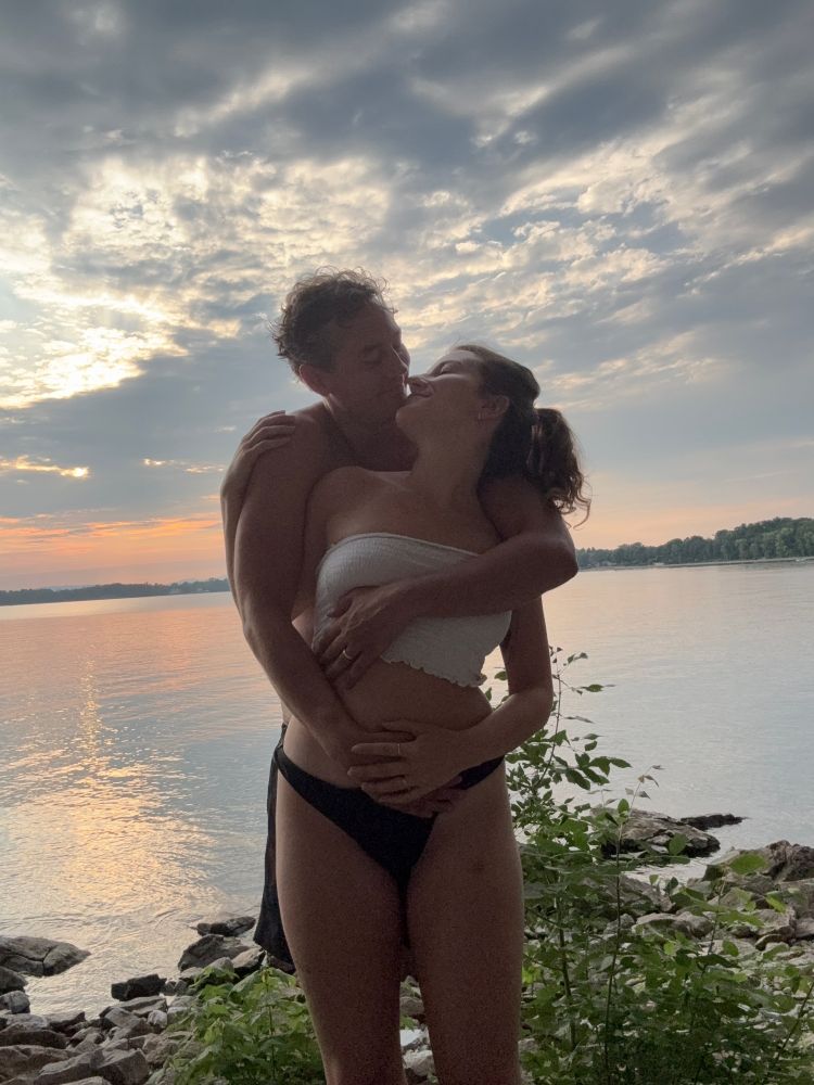 A husband and wife, madly in love, embrace in front of a sunset on a lake 