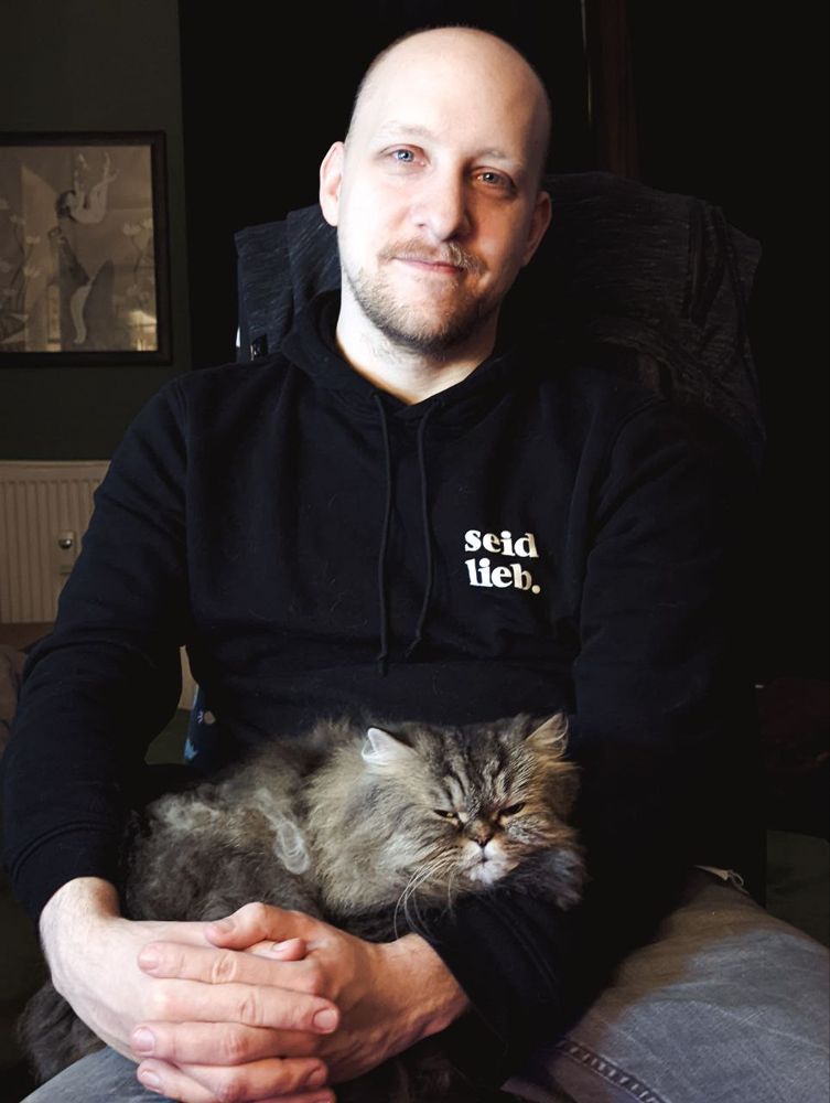 Ein lächelnder Mann mit Glatze (Henrik) sitzt auf einem Stuhl, er trägt einen "SEID LIEB" Pulli und hat eine wuschelige Katze aauf dem Schoß.