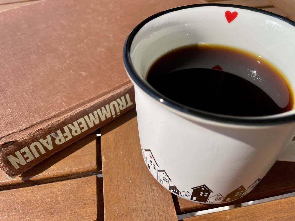 Kaffeebecher auf Balkontisch in der Sonne, daneben ein Buch mit dem Wort „Trümmerfrauen“ auf dem Buchrücken