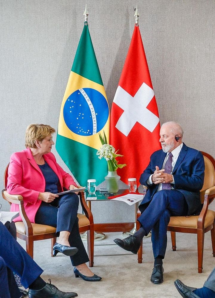 O presidente Lula conversa com a presidenta do Conselho Federal da Suíça, Viola Amherd. Ao fundo, as bandeiras do Brasil e da Suíça.