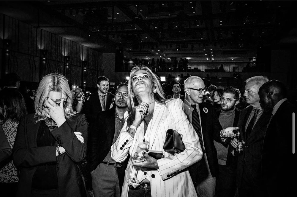 Photo of the crowd watching the election results with upset Luhks. On the left, a woman looks down while massaging her temples with one hand. In the middle, a woman looks up at the screen with one hand raised towards her face and a wine glass in the other, securing an expensive clutch in the crook of her elbow. In the background on the right, an elderly man wearing glasses hold out his left hand, palm up, while speaking to three other men in suits with a concerned look on his face. 
