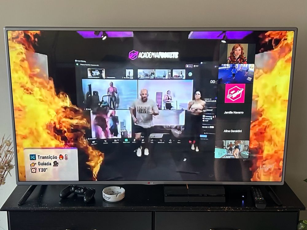 A TV with a live workout session. A man and a woman are running in place in front of a giant projection of the Zoom gallery showing of people working out at home. On either side of the screen, giant columns of fire erupt from below. 