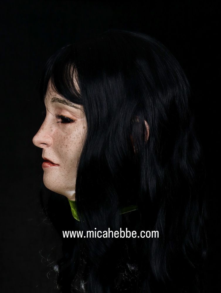 Jenny mask against a black background. She is pale with freckles and long dark hair. She had a green velvet ribbon around her neck. Side view 