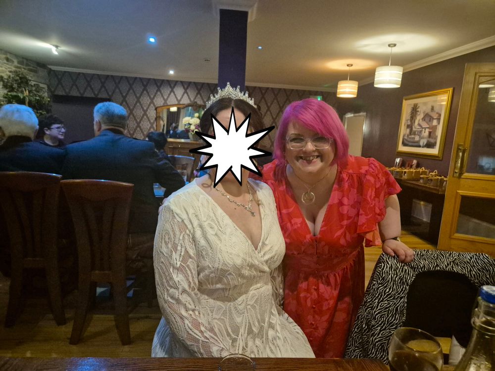 The bride is sitting in a chair in a hotel function room, there is a table of people behind her. She is wearing a long sleeved white lace dress and a silver tiara. Dawn is leaning forwards beside her, wearing a bright red and hot pink floral dress and her hair is bright pink.