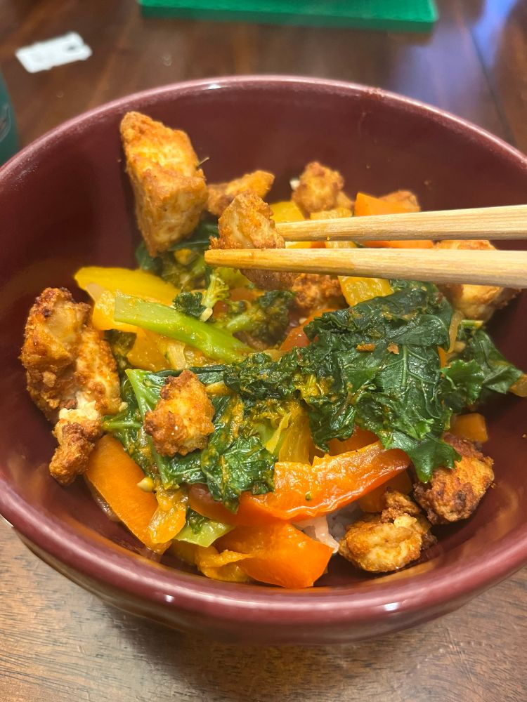 Closeup of a veggie curry with chopsticks holding a piece of air fried tofu