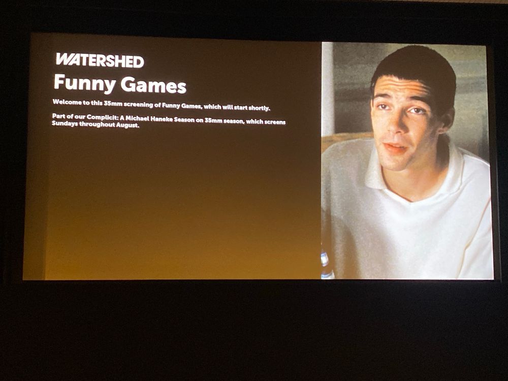 A cinema screen showing the intro card for the film Funny Games. A creepy guy in whites in on the right hand side.