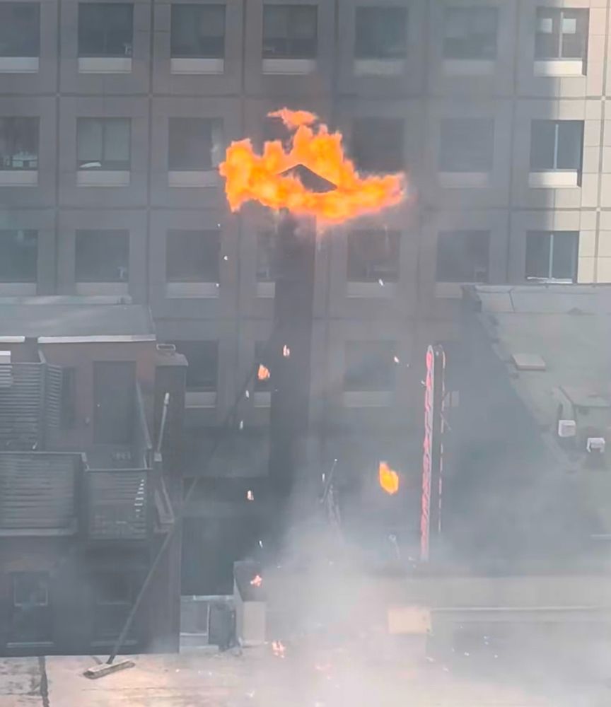 A rooftop chimney against the backdrop of an office building. The crown of the chimney is wreathed in orange flame, and white smoke gathers at its base where it protrudes from the roof