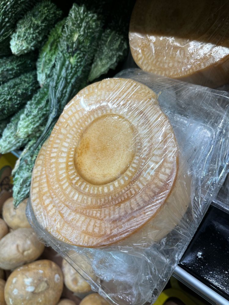 Photo of a cross section of a banana stem wrapped in thin plastic and surrounded by many tightly wrapped banana leaf stalks surrounded by other vegetables in Asian food market.   The banana piece is beige to brown, with a center circular stem, evenly yellow. Around it are thin halfmoon-shaped cross sections of leaf stalks, tightly wrapped around the central stem. Each leaf part shows banded patterns in beige and white. Own photo, (c) Lena Struwe, 2024. 
Musa sp. , Musaceae. 