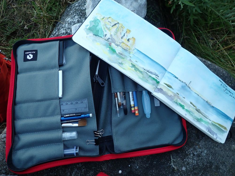 My field kit for nature journaling outdoors. Photo of red zippered bag, gray on inside with many compartments with 2 small watercolor palettes (businesscard-sized in metal), ruler, brushes, pens and pencils, tiny water spray bottle, waterbrush, and metal clips.  On top of that is the sketchbook with a watercolor image of a long, horizontal landscape from the coast of Gotland - blue sea along the horizon, light blue sky, beige and yellow cliffs to the very left, and in the distance a narrow sliver of an island with a thin lighthouse on it; in the foreground along the whole double page are the beach with green (several shades) of shrubs and herbs, some gray low rocks along the shore, and a sandy beach in the center. The sketching kit and sketchbook is laying on some rocks among grass.  (Sketch kit is made by Art Toolkit). Photo and art by (c) Lena Struwe, 2022. #naturejournaling #watercolor #plein air