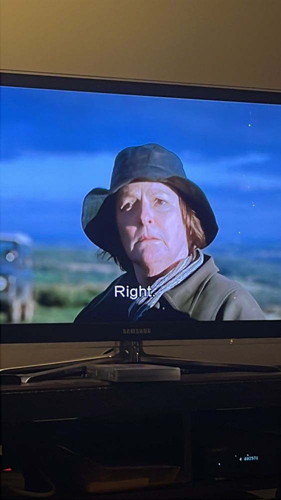 Photo of detective Vera Stanhope, middle-age white woman in green field hat, olive green raincoat, and a scarf, short brownish hair, behind her blue sky and blurry green lands - as shown on TV screen. On the photo is also the subtitle ‘Right’ in white text. 