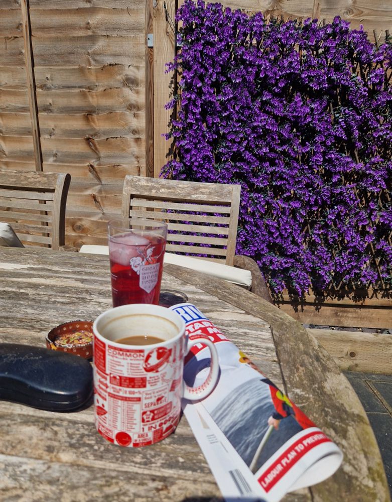 Coffee, sunglasses case, pint of grenadine squash with giant ice cube, Private Eye. On a wooden garden table with a chair and a fence in the background 