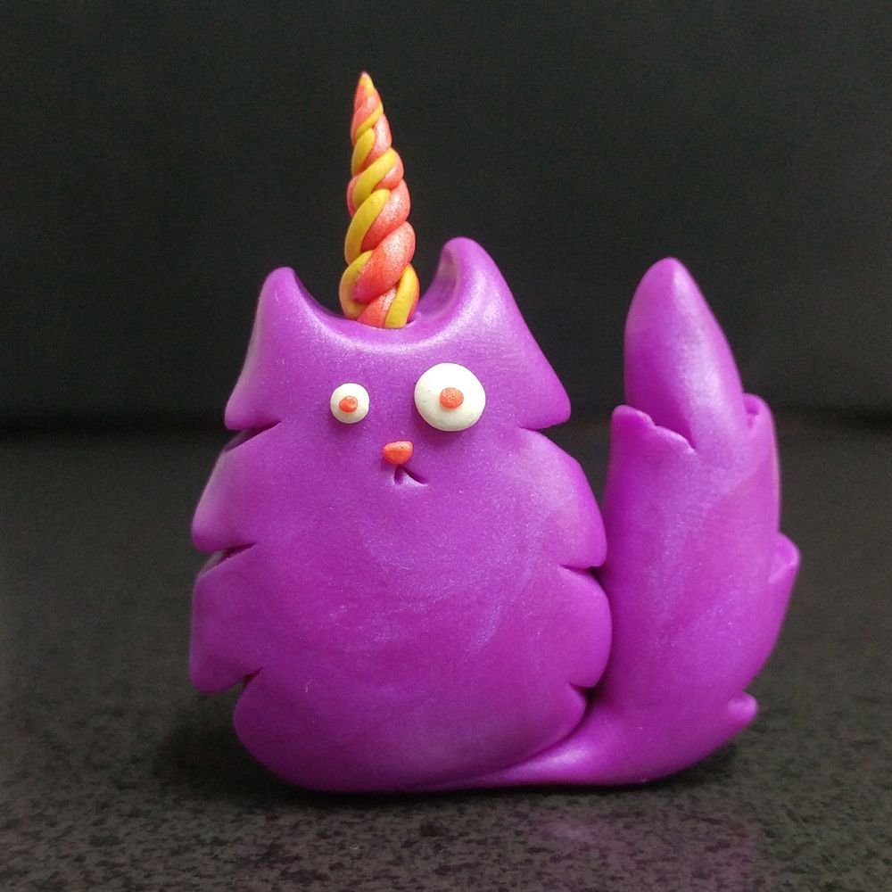 Photo of a fluffy-looking purple, polymer clay cat with a yellow and pink unicorn horn