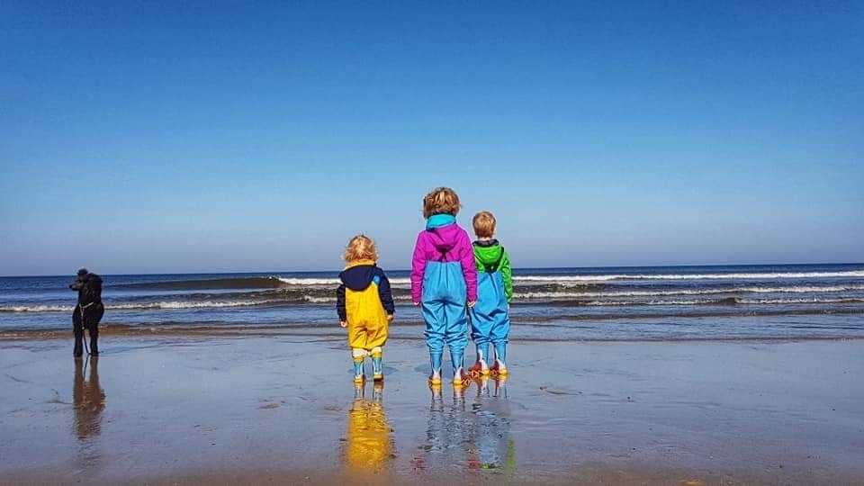 3 Kinder in bunten Wasserhosen und Stiefeln stehen mit einem schwarzen Pudel am Strand und schauen auf das Meer. Der Himmel ist blau und die Wellen tragen kleine Schaumkronen.