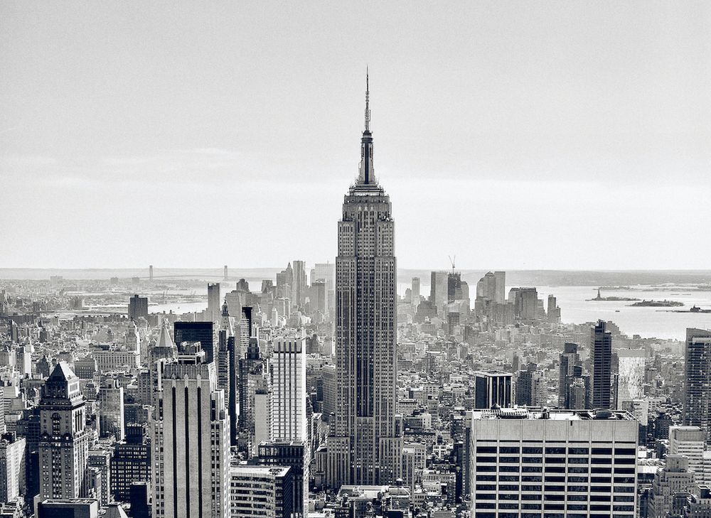 an expansive view from 30 Rockefeller plaza rooftop to the empire state in the foreground and then south all the way to the Verrazzano-Narrows Bridge just visible on the left just below the horizon and the statue of liberty and Ellis Island on the right