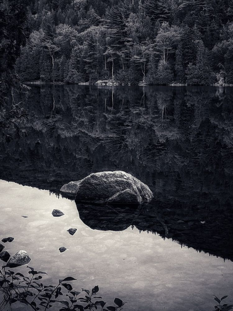 a monochromatic image of a sunlit rock in the shallows just at the boundary of the bright reflected and overcast sky and the dark, shadowed reflection of the forested hill on the opposite bank creating a smooth and perfect mirror