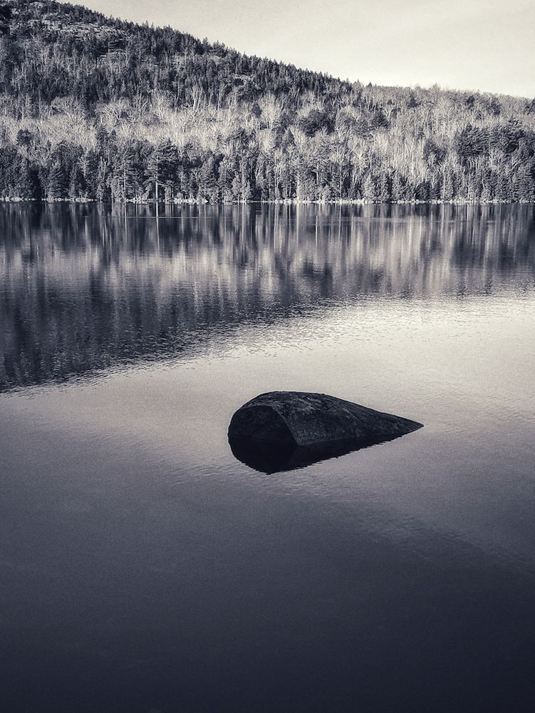 an image of a seemingly floating stone, dark in foreground shadow, in placid, shallow waters with the blurred reflection of the opposite shore in late fall foliage on the horizon