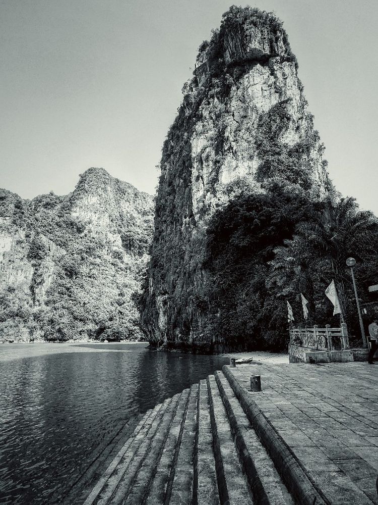 a stone stairway ascends from the water onto a terrace entry way onto a small island beach dominated by an impressive karst tower and mountains in the distance