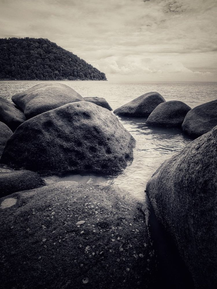 A collection of boulders, closely grouped, bathe in the warm shallows waters at the edge of monkey beach, in the distance the horizon and a small peninsula at the opposite end of the crescent shaped bay