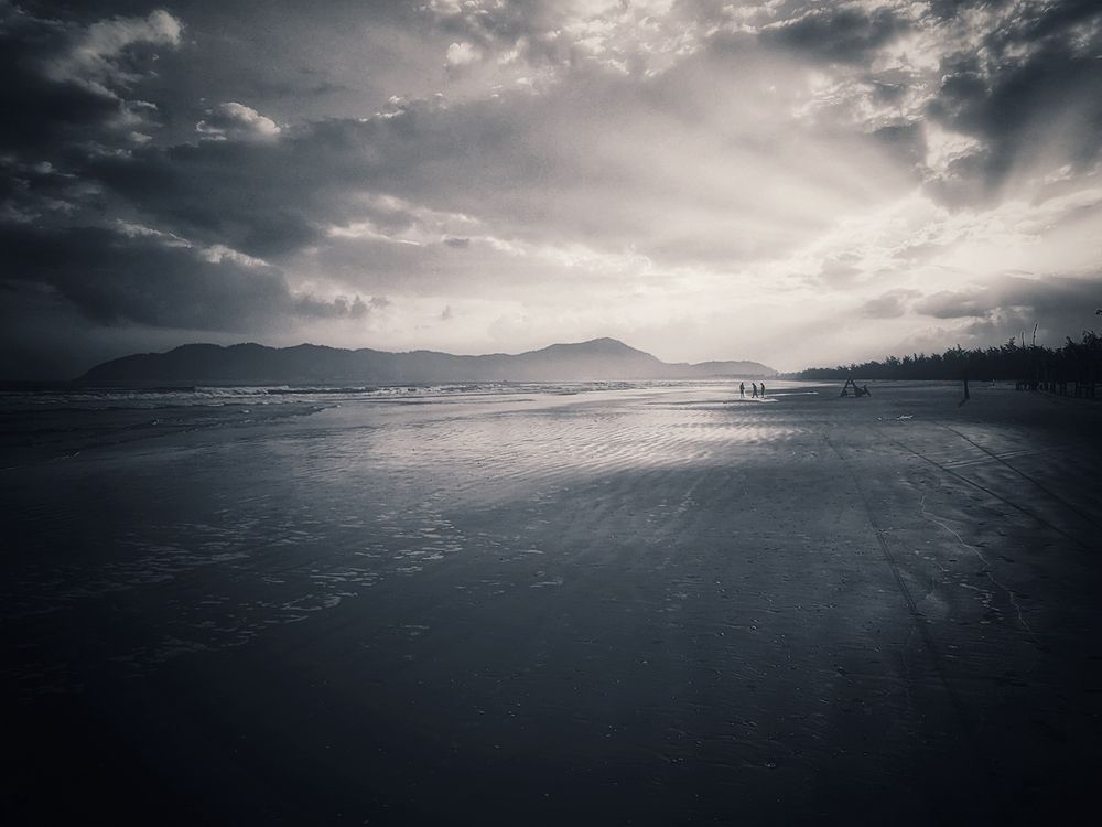 dramatic morning light rays breaking through the heavy storm clouds onto a dark and nearly deserted beach, in the distance a group of female fishermen pull in a large net by hand... it is indeed back breaking work which they perform for hours every morning with authentic smiles