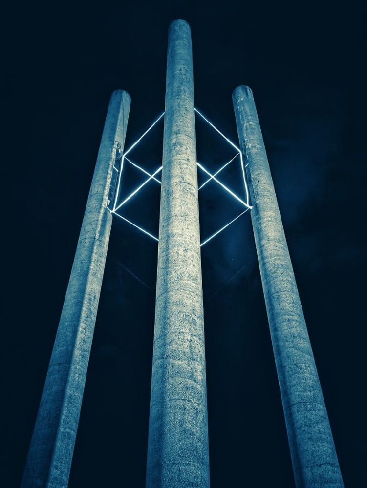 3 visible steel blue pillars (4th pillar can't be seen) rise into the night sky, up near the top a light cube made of electrical wiring and a lovely Sci Fi element at night