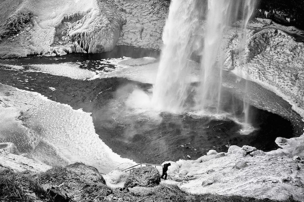 a view from above the large plunge pool of a waterfall in winter - the spray from the waterfall has covered the ground and then frozen into nodules making the terrain extremely dangerous, below a sole figure climbs the stairs to the walking path that will take you into a partial cave with a view from behind the curtain