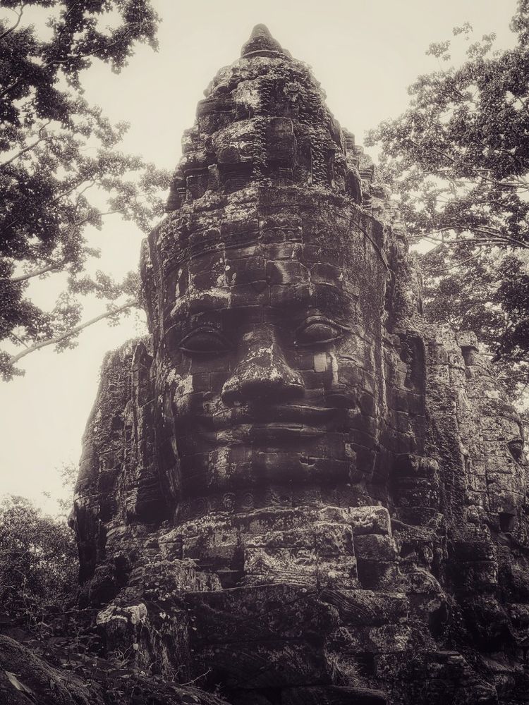 a giant stone-carved and weather worn face of a gatekeeper rises from the jungle overgrowth against a blazing and humid summer sky