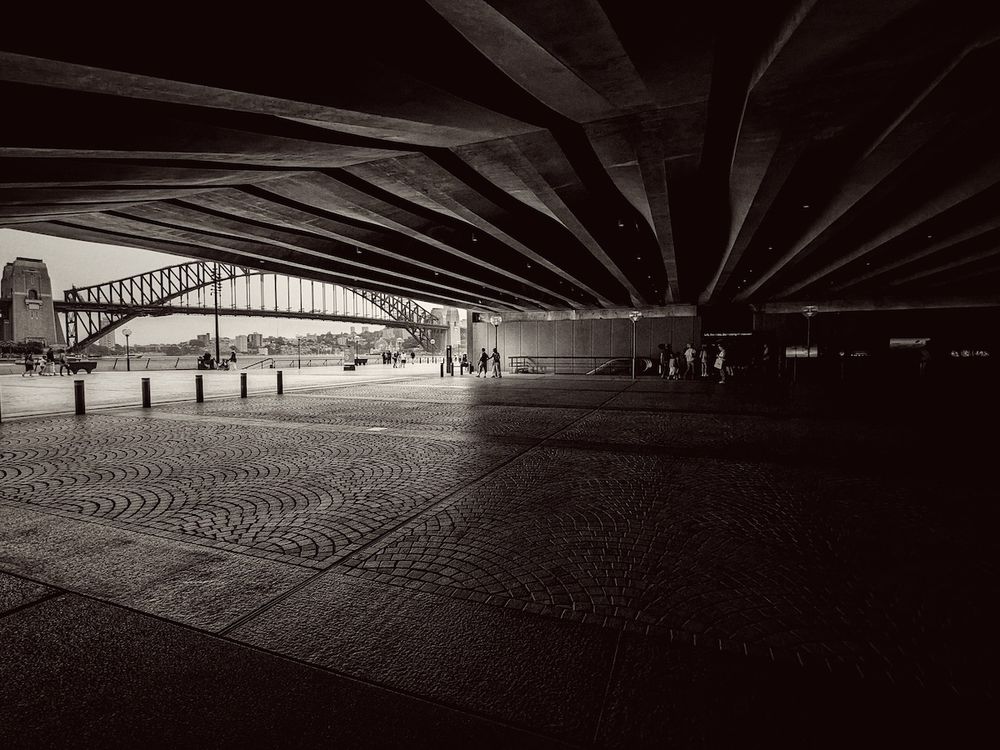 an image of handsome beams of arched concrete, the dim but impressive underworld beneath the sydney opera house, lit by the bright daylight reality of midday, in the distance the sydney harbor bridge is visible