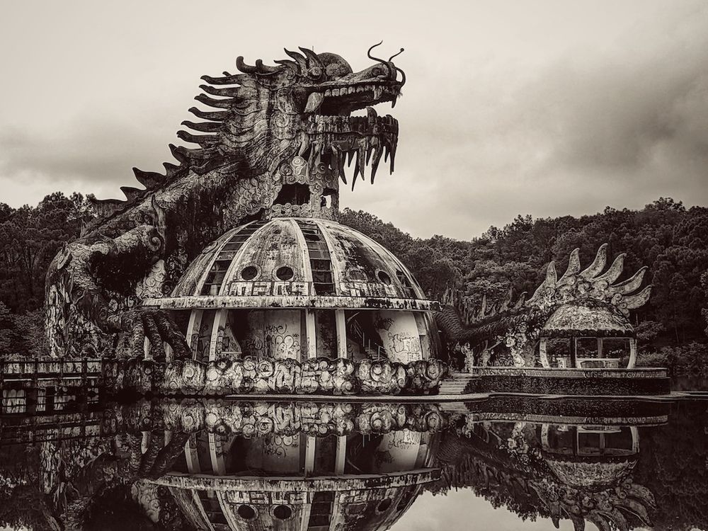 an image of a once spectacular structure of a dragon perched on semi spherical base placed strategically in a circular pond, hidden in the remote jungle. this forgotten relic is located in an abandoned water park -- and you can climb up and through the dragon to a viewing platform in the space between the upper and lower jaws