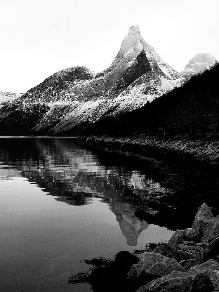 a high contrast wintry image of Norway's national mountain with a thin coating of ice and against a sky of white icy mists, as it slopes down to the pristine fjord below and the tranquil water's edge