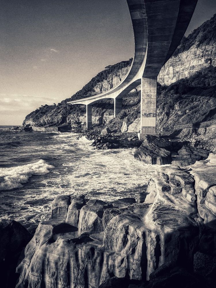 the view taken from beneath the Sea Cliff Bridge on a rocky ledge looking down at the crashing waves, the arch of the sleek concrete bridge curving gracefully above 