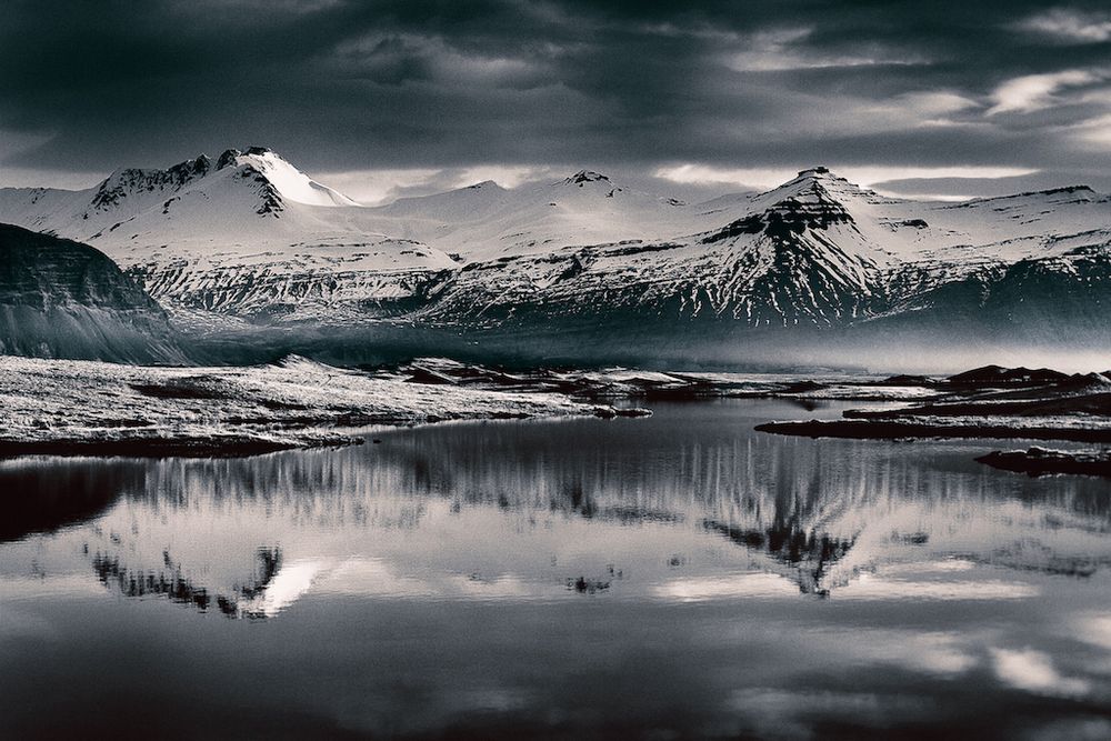 a coastal mountain ridge covered in snow and ice is softly reflected in a deep pool of runoff that is slowly making its way to the sea just out of sight to the right, dark clouds float in the sky behind the mountaintops 