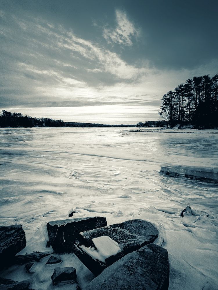 an image of the frozen 'wasteland' of the Harraseeket River delta taken from the icy shoreline strewn with large boulders, the chunky ice extends far into the distance where the opposite shoreline is a thin strip of shadow, above a cold and fiery sky  