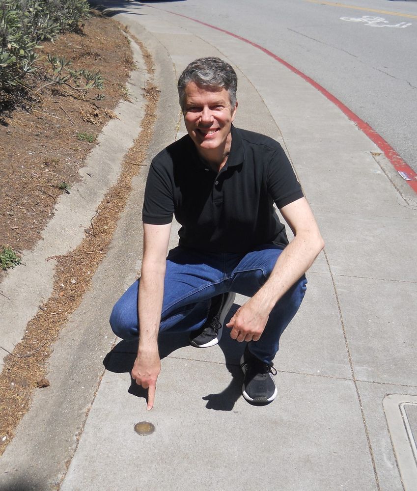 Man squatting and pointing to a brass disk in a sidewalk.