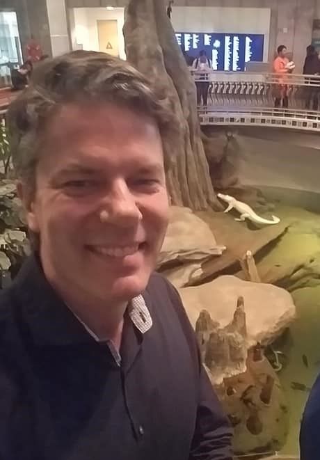 Smiling man in front of an exhibit at an aquarium featuring an albino alligator. 