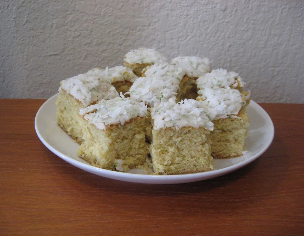 White plate with small slices of a yellowish cake with white glaze and coconut on top, on a wooden table against a white wall.