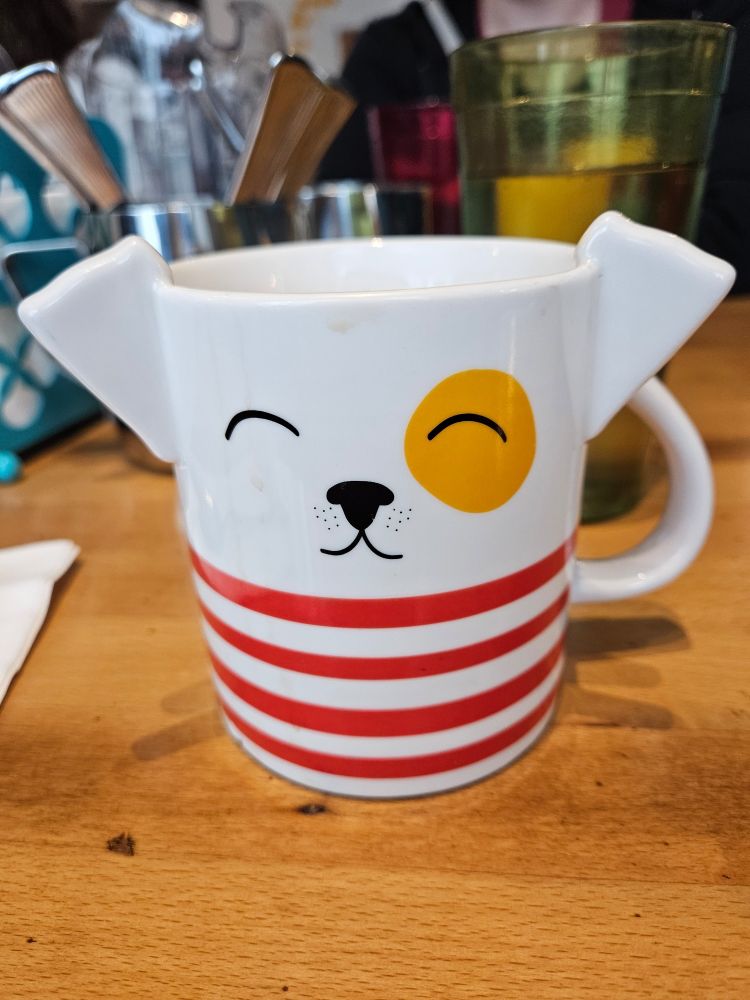 A picture of a coffee mug with a smiling cartoon dog face with a yellow spot over one eye. The mug has dog ears. The bottom half of the mug is decorated with red stripes.