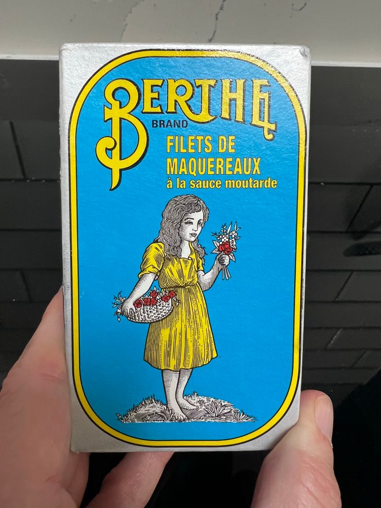 A tin of mackerel, wrapped in cerulean blue paper with an early 20th style drawing of a girl holding flowers and wearing a yellow dress. The yellow filigree writing says ‘Berthe brand filet de maquereaux’